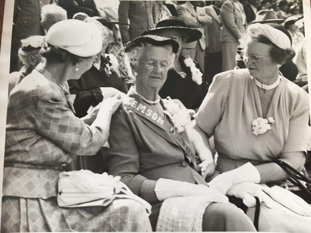 Black and white photo from 1954 of three women dressed formally. The woman in the middle is wearing a sash.