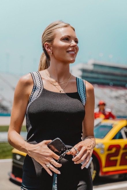 A woman in a black top holds a lanyard and phone, and a yellow and red race car sits in the background behind her.