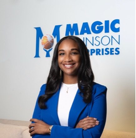 A woman in a white top and royal blue blazer poses in front of a partially blocked sign that says Magic Johnson Enterprises. 