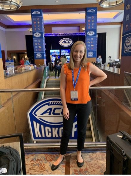 A woman in black dress pants and an orange tops poses in front of a sign that says ACC Kickoff. 