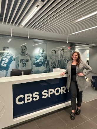 A woman in a black top, black pants and a gray blazer poses in front of a desk that has a CBS Sports sign on its front. 