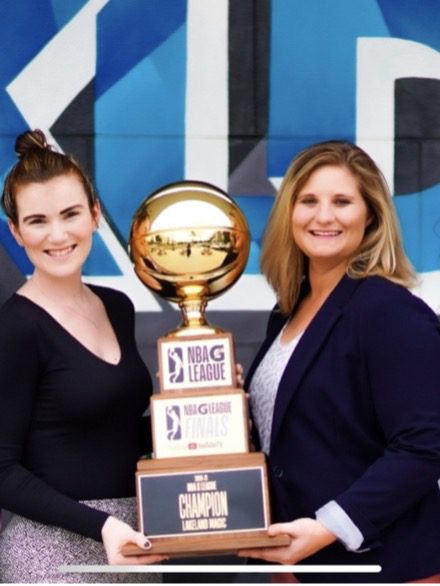A woman in a black top and purple floral-patterned pants and a woman in a top and navy blue blazer together hold an NBA G League championship trophy. 