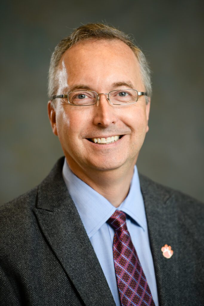 A head and shoulders studio photo of a man, John DesJardins, who is smiling. He is wearing small framed glasses, coat, tie and a Clemson University Tiger Paw pin on the lapel of his coat.