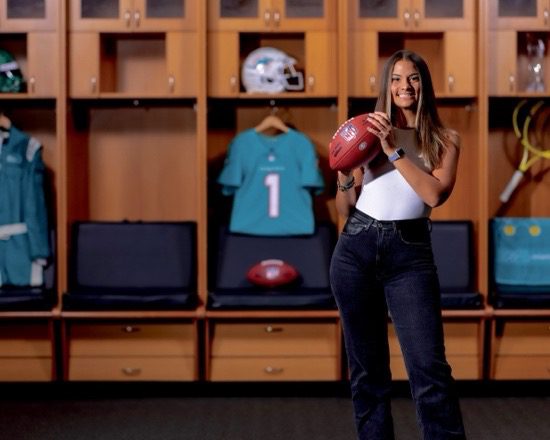 A woman in jeans and a white sleeveless top holds a football inside of a football locker room with a football, teal football jersey and white Miami Dolphins football helmet in the background. 