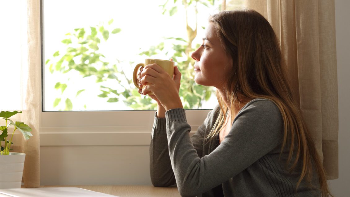 Side view of a relaxed woman looking through a window sitting in a table and holding a cup of coffee at home