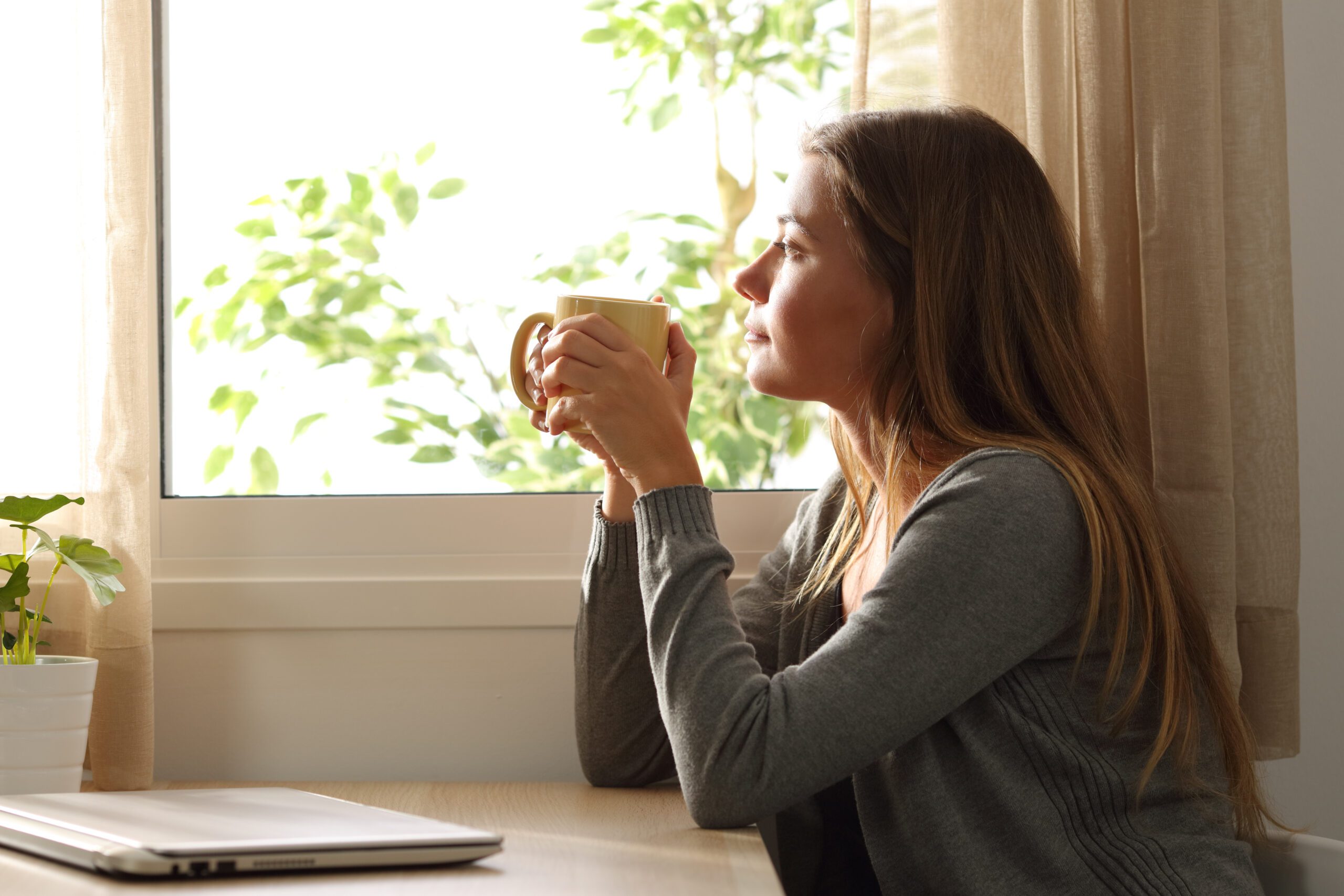 Side view of a relaxed woman looking through a window sitting in a table and holding a cup of coffee at home