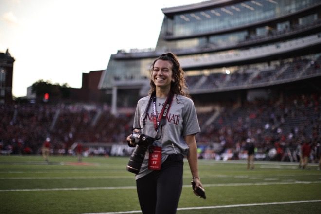 A woman wearing a gray T-shirt that says Cincinnati and black jeans holds a digital camera around her neck while standing on a football field. 