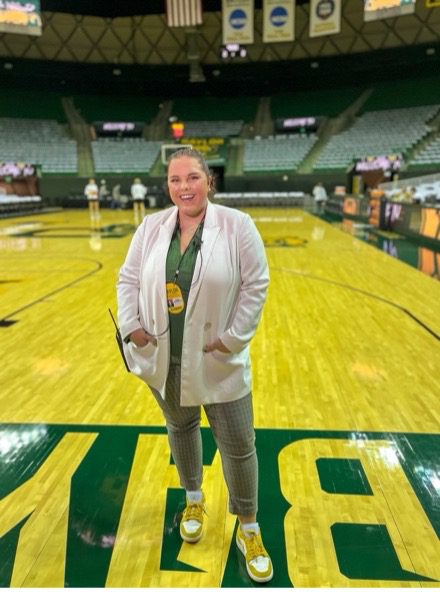 A woman in gray pants, a green top and a cream-colored cardigan stands on a green and neon yellow court floor inside of the Baylor basketball arena. 