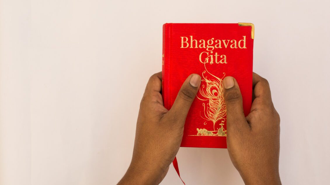 A pair of hands holds a small red copy of the book Bhagavad Gita in front of a white background.