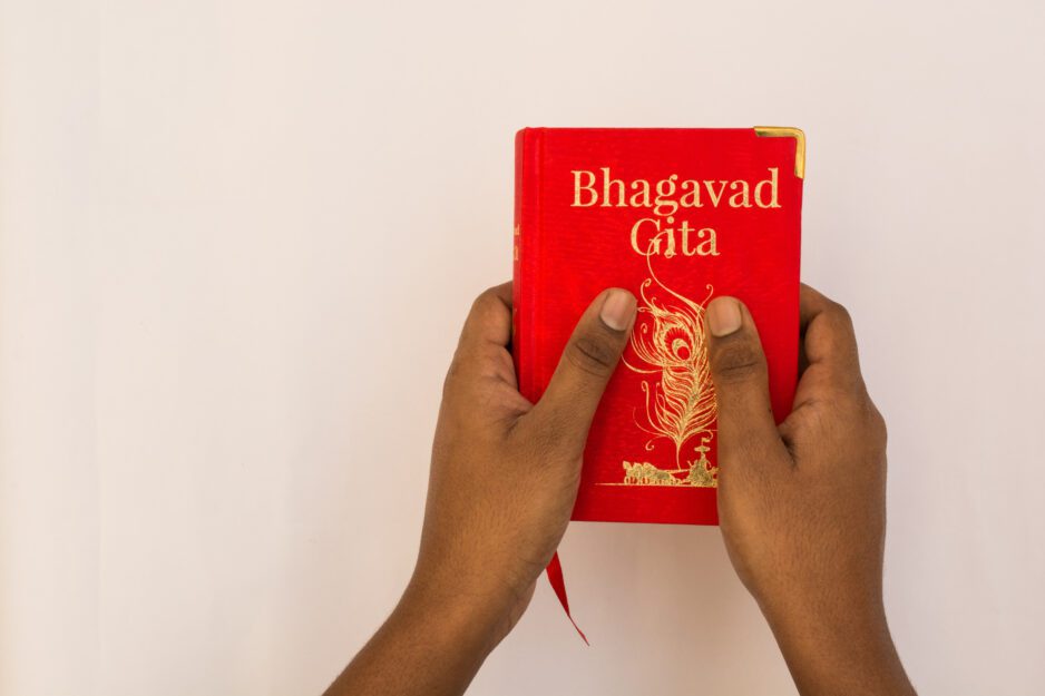 A pair of hands holds a small red copy of the book Bhagavad Gita in front of a white background.