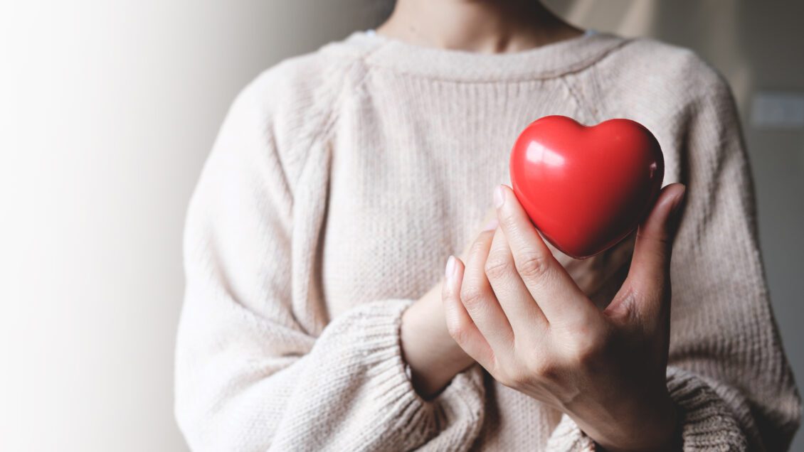 A woman in a tan sweater holds a red toy heart in her hands in front of her chest.