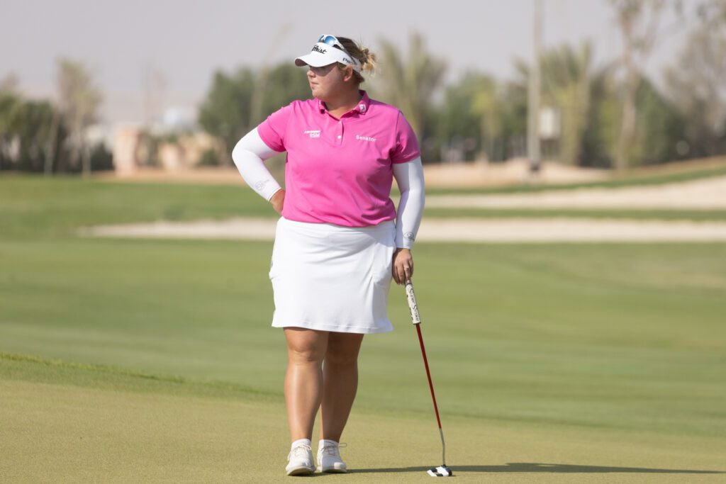 A woman wearing a white skirt, a white long-sleeved top layered with a pink polo and a white visor stands on the green of a golf course and holds a golf club. 