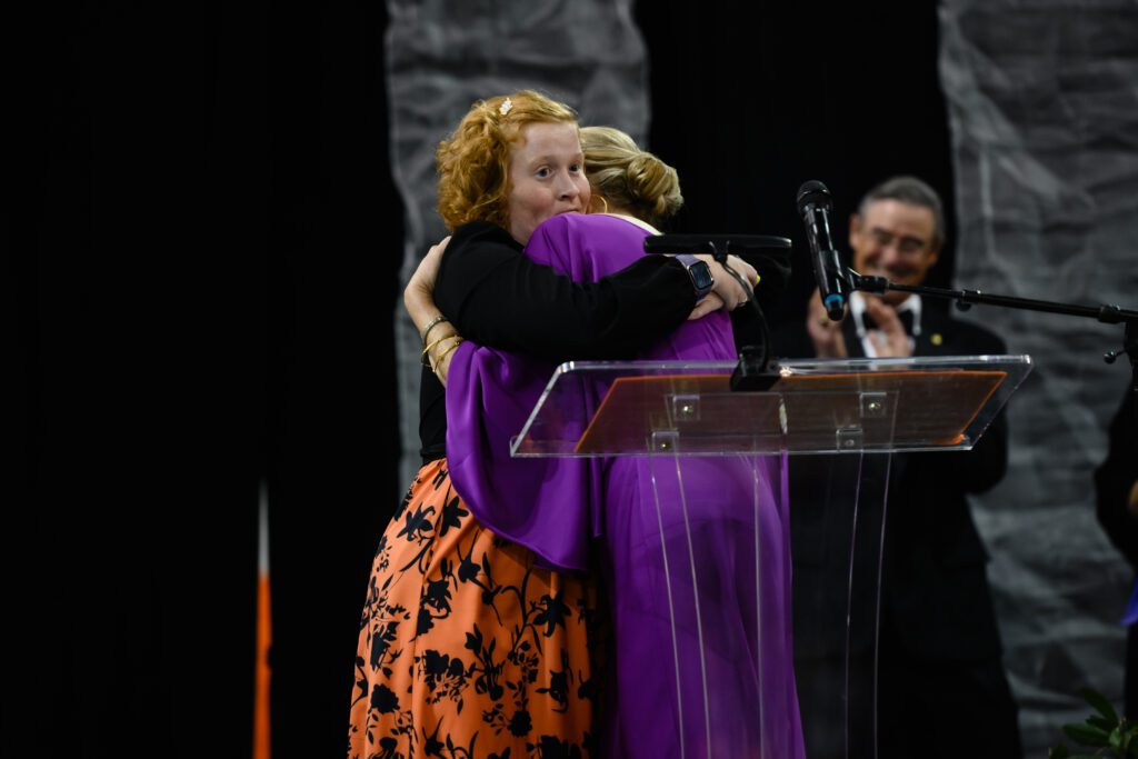 A red-haired young woman with her hair pulled back wears an orange and black dress and embraces a woman wearing a purple ballgown while a man claps in the background. They are standing on a stage in front of a podium and microphone. 