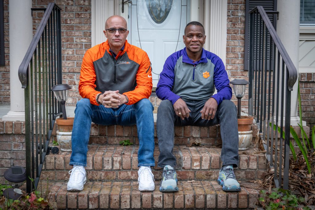 Two men wearing jeans and Clemson-branded jackets sit on the front brick steps of a residential home. 