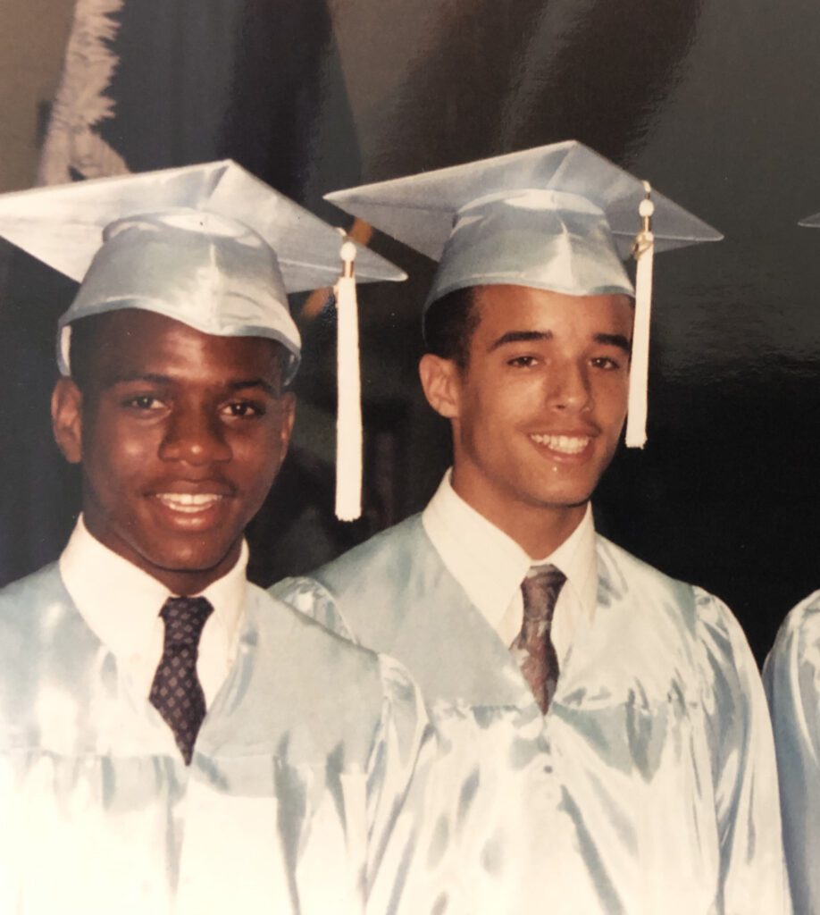 Two high school male students wear light gray caps and gowns, white shirts and brown ties at a graduation ceremony. 