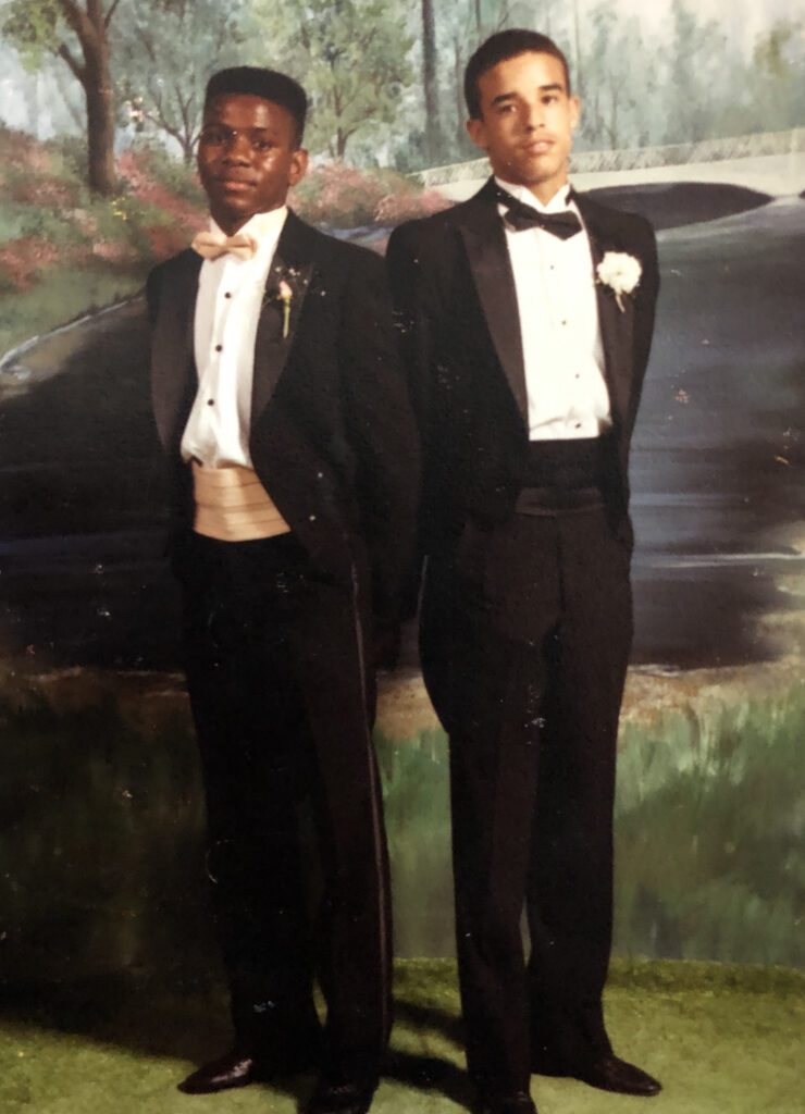 Two high school male students wear tuxedos while posing in front of a nature scene backdrop. 