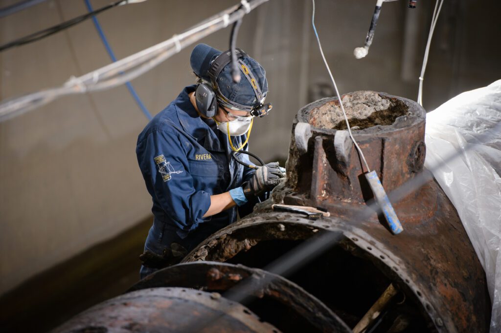 A man wearing a mask and a blue uniform and blue ball cap turned backward is holding a tool beside the metal shell of a recovered submarine, the H.L. Hunley