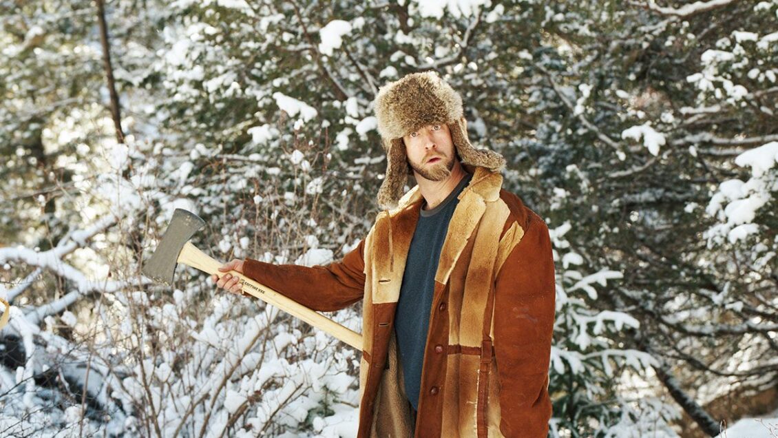 Josh Blue looks confounded while holding an axe in a snowy winter setting.