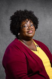 A studio headshot of an African American woman with short, curly hair wearing glasses and a yellow v-neck shirt and burgundy cardigan sweater. 