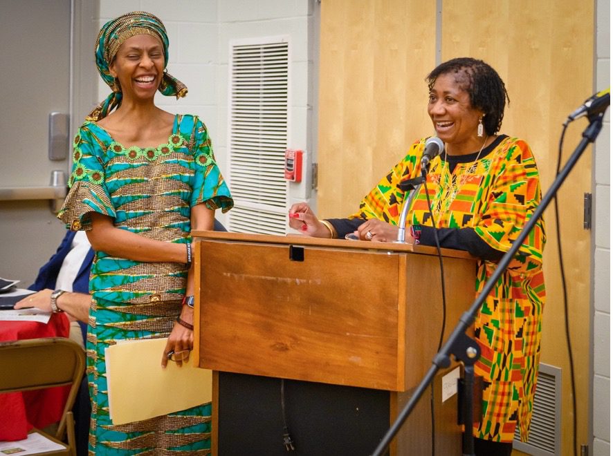 Two Black women in traditional African attire smile as they stand around a lectern.