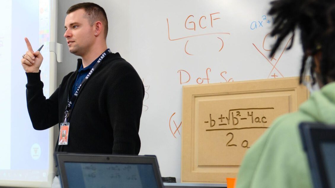 A male student teacher wearing a black quarter-zip sweater and khakis and a female teacher wearing a black dress and purple cardigan stand in front of a projector screen in a high school classroom.
