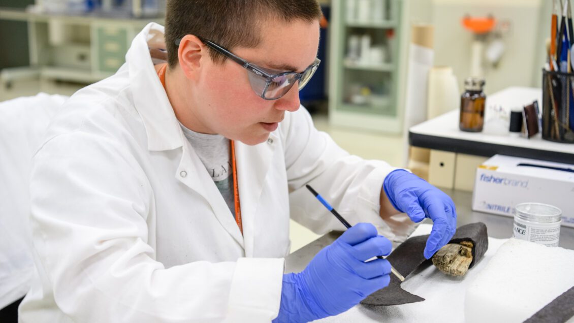 A student wearing a white lab coat, blue gloves and safety glasses uses a pen-like instrument to inspect a piece of submarine metal.