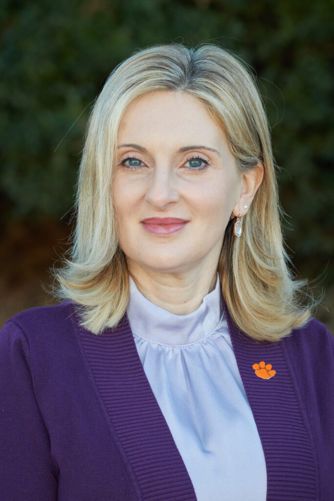 Woman (Karolina Mukhtar) with shoulder length hair, parted in the center, wearing a blazer, blouse with a mock neck, and a Tiger Paw pendent on her blazer lapel.