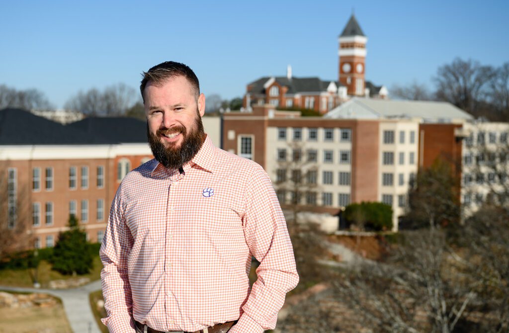 A bearded man wearing a orange Clemson Oxford shirt standing on a rooftop smiling.