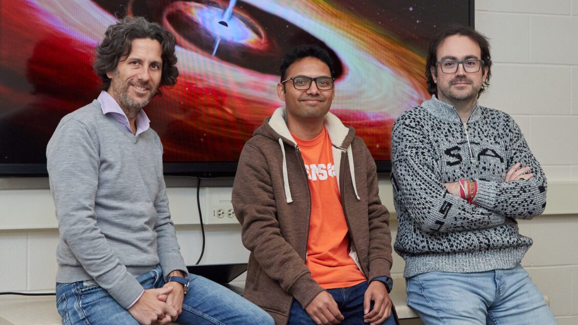 Three men are sitting in front of a photo of a supermassive black hole