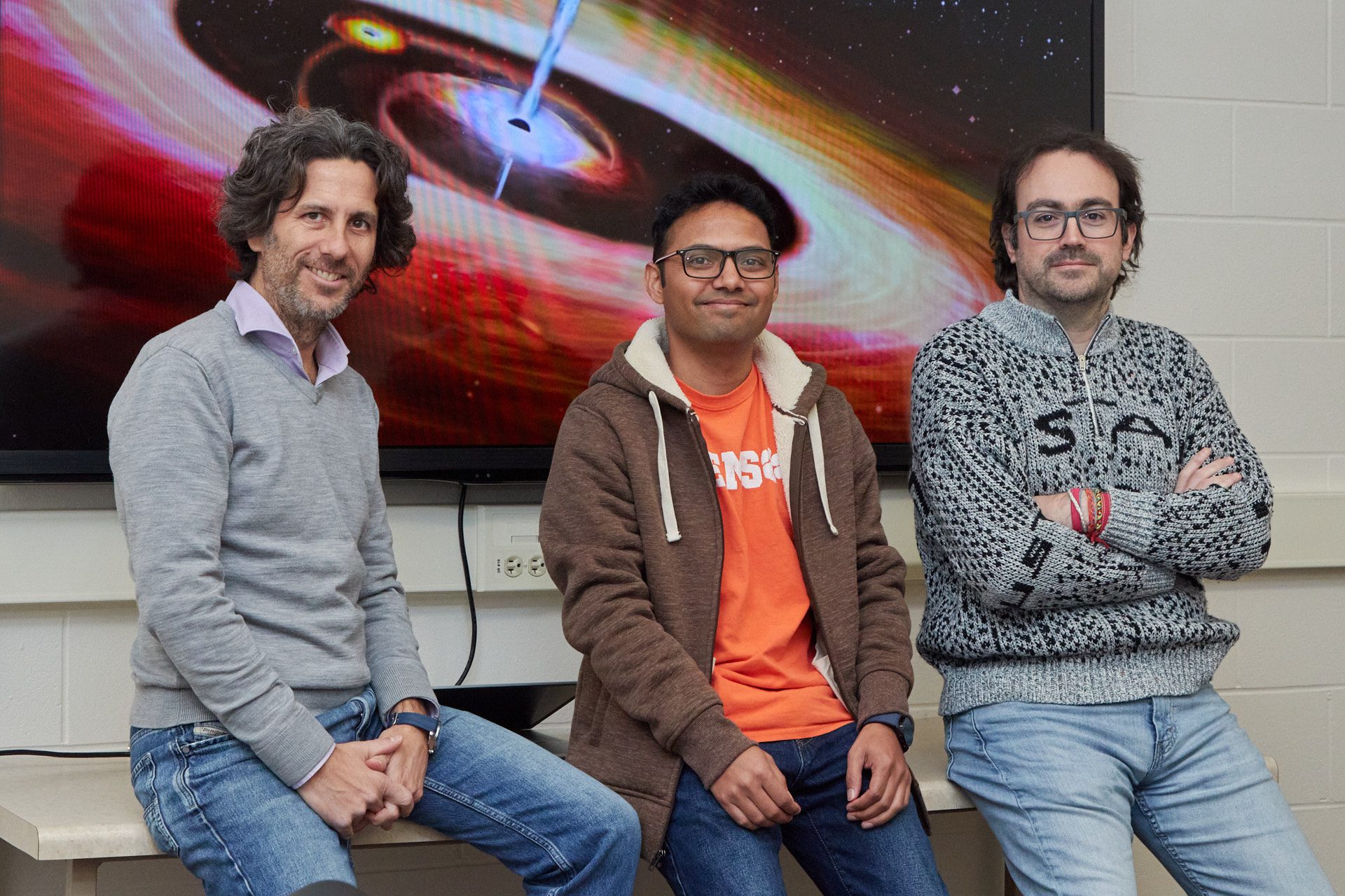 Three men are sitting in front of a photo of a supermassive black hole