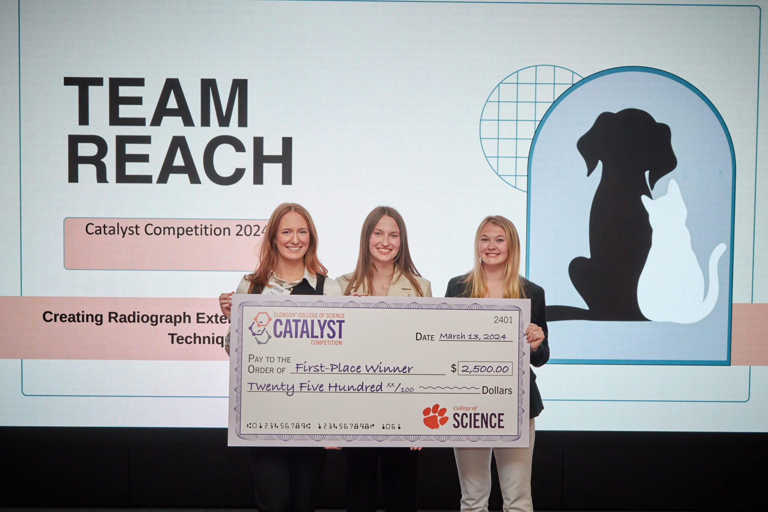 Three women holding a big check for winning the College of Science Catalyst Competion stand in front of a screen with the name of their team and an outline of a dog and cat