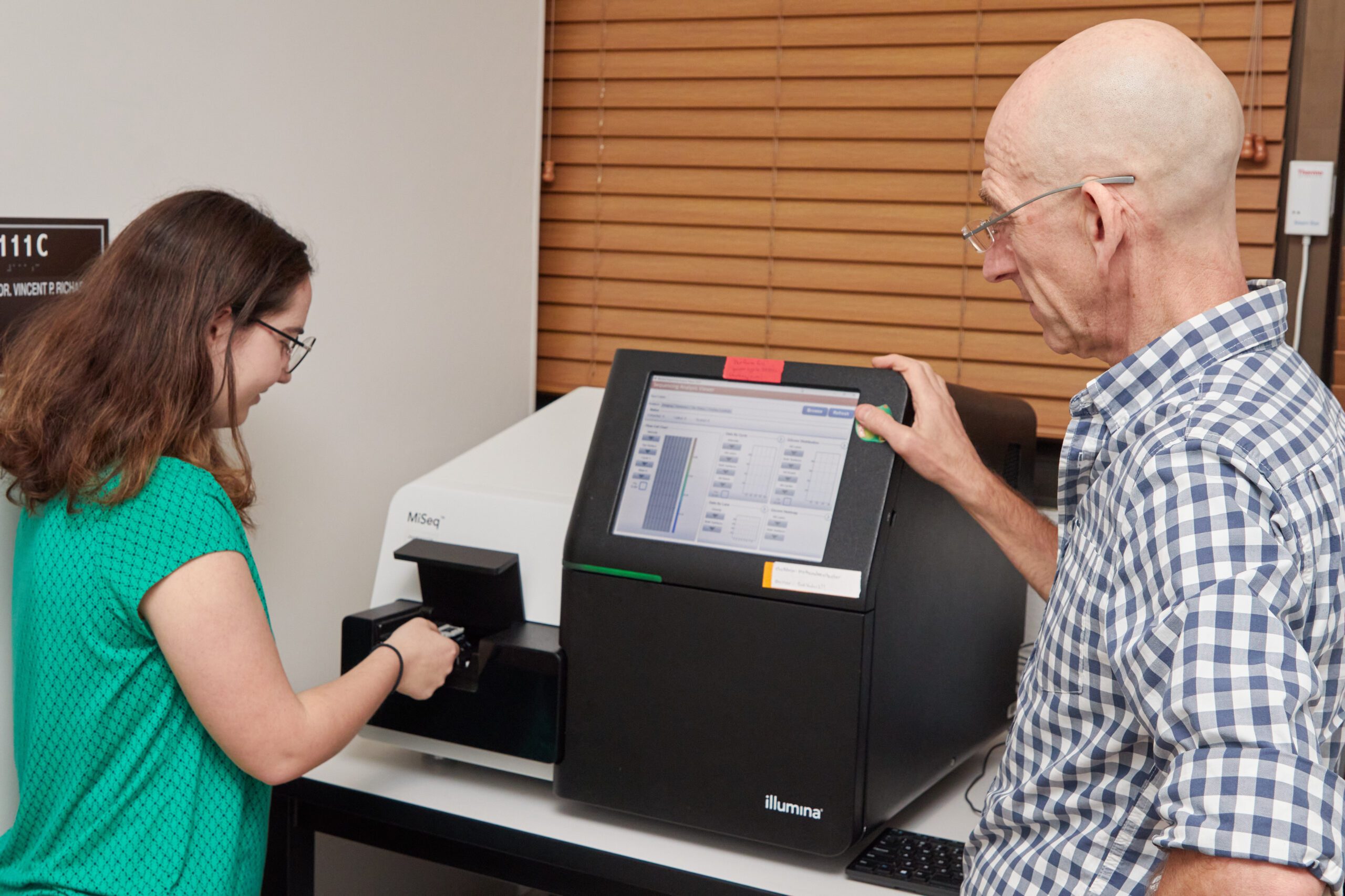 A woman wearing a green shirt is inserting something into a sequencing machine while a man watches.