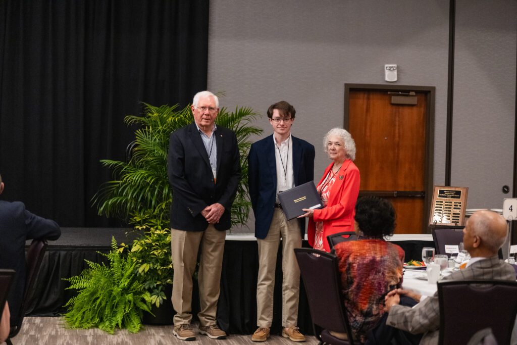 A male student dressed in a blazer is photographed receiving an award from two faculty.