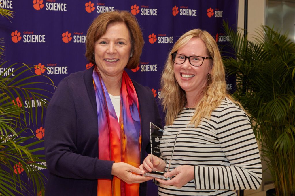 Two women standing in front of a College of Science backdrop with one holding a trophy