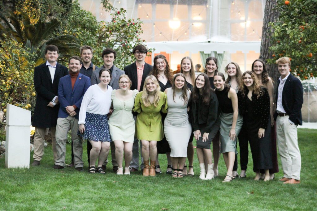 A group of 18 well-dressed young people smile on a lush lawn with hanging lights and orange trees. Katherine Harland is on the second row.