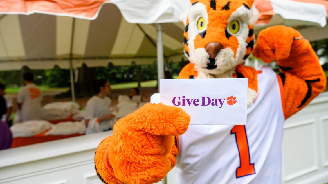 Clemson Tiger mascot holds sign that reads Give Day. He is standing in front of an outdoor tent.
