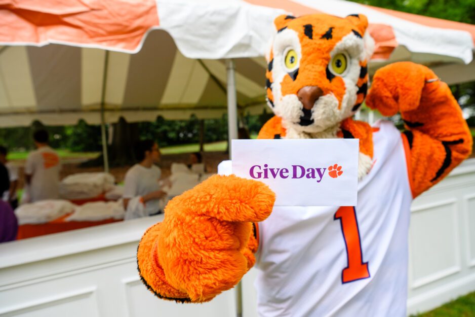 Clemson Tiger mascot holds sign that reads Give Day. He is standing in front of an outdoor tent.