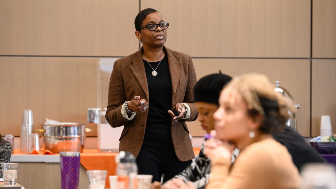 An African American woman wearing black-rimmed glasses and a brown blazer presents at the front of a room.