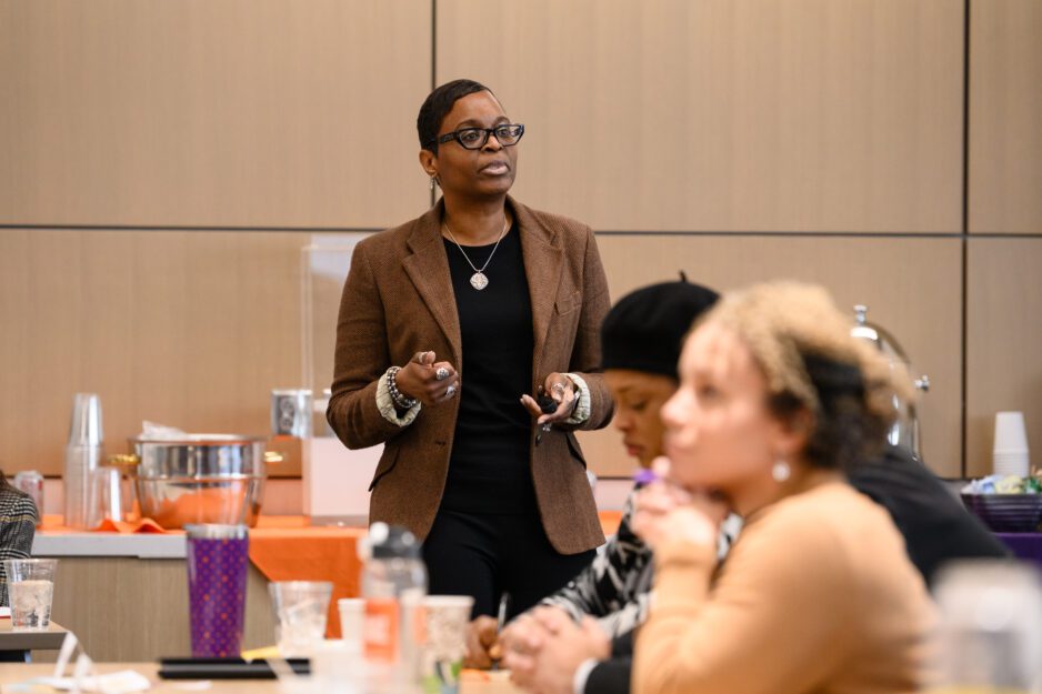 An African American woman wearing black-rimmed glasses and a brown blazer presents at the front of a room.