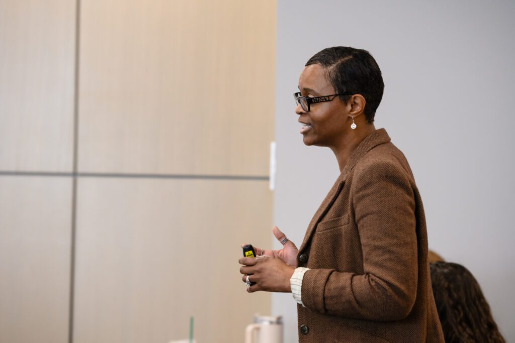 An African-American woman stands at the front of a room. She is wearing a brown blazer and black glasses.