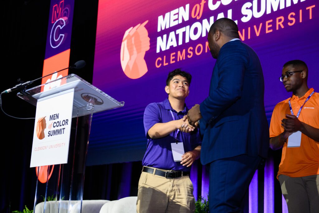 A young, latino male wearing a short-sleeve collared purple shirt shakes hands with an African American man wearing a blue suit. They are standing on a stage with signage that reads "Men of Color."