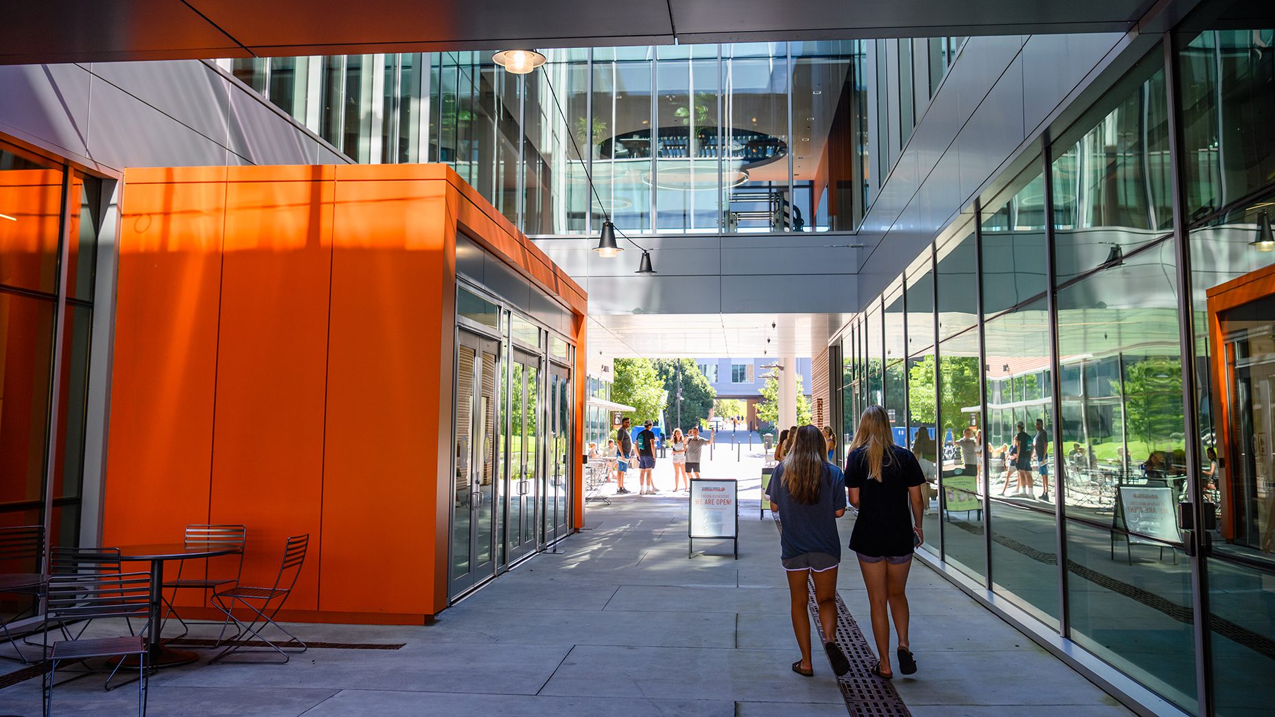 Two women walk through an outdoor corridor lined with glass and an orange wall