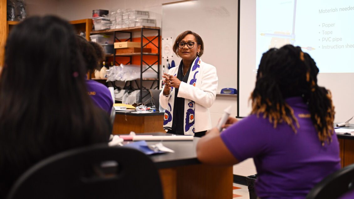 A Black woman stands in front of two seated Black female students, both wearing purple shirts, and she is presenting something to them inside a classroom laboratory.