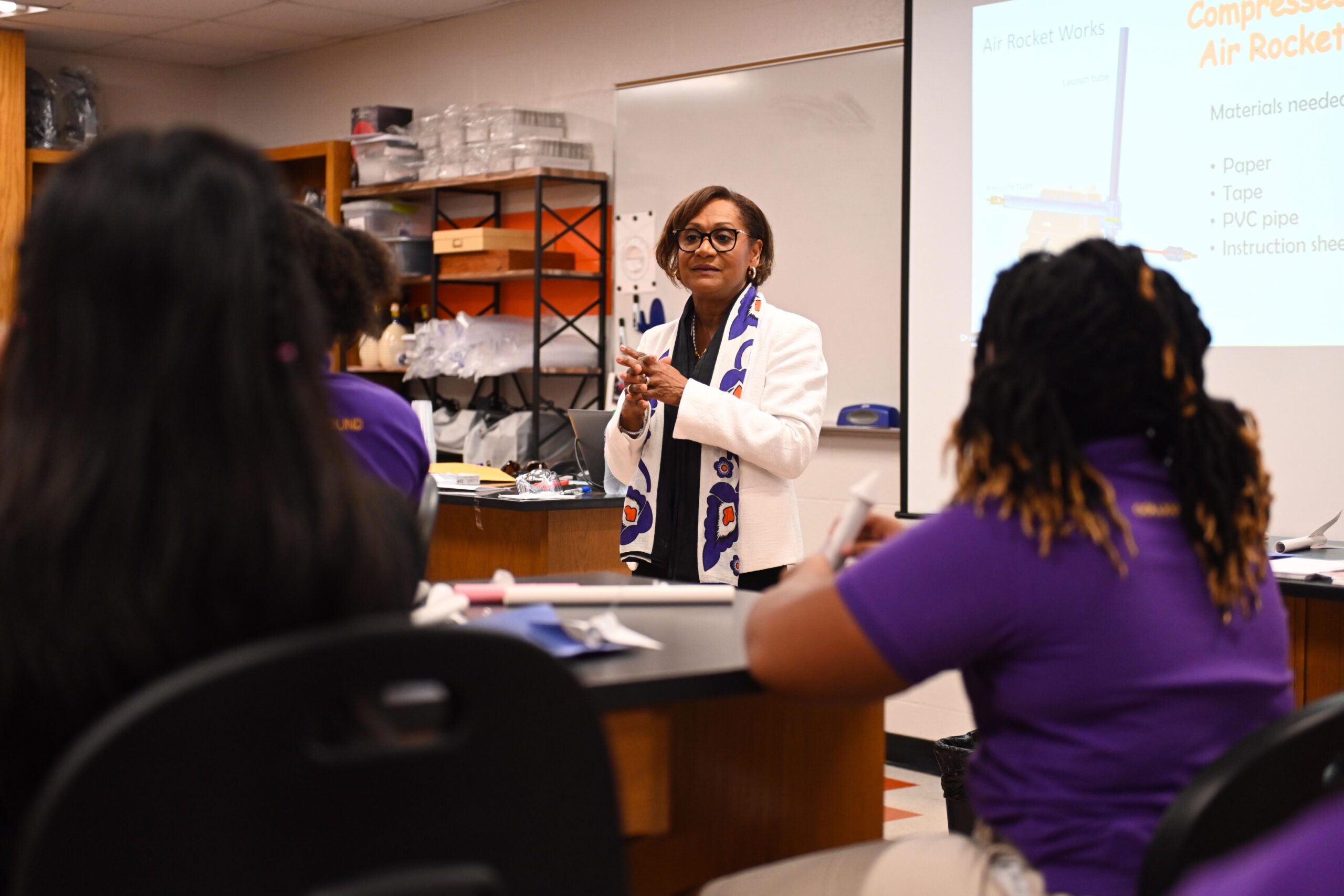A Black woman stands in front of two seated Black female students, both wearing purple shirts, and she is presenting something to them inside a classroom laboratory.