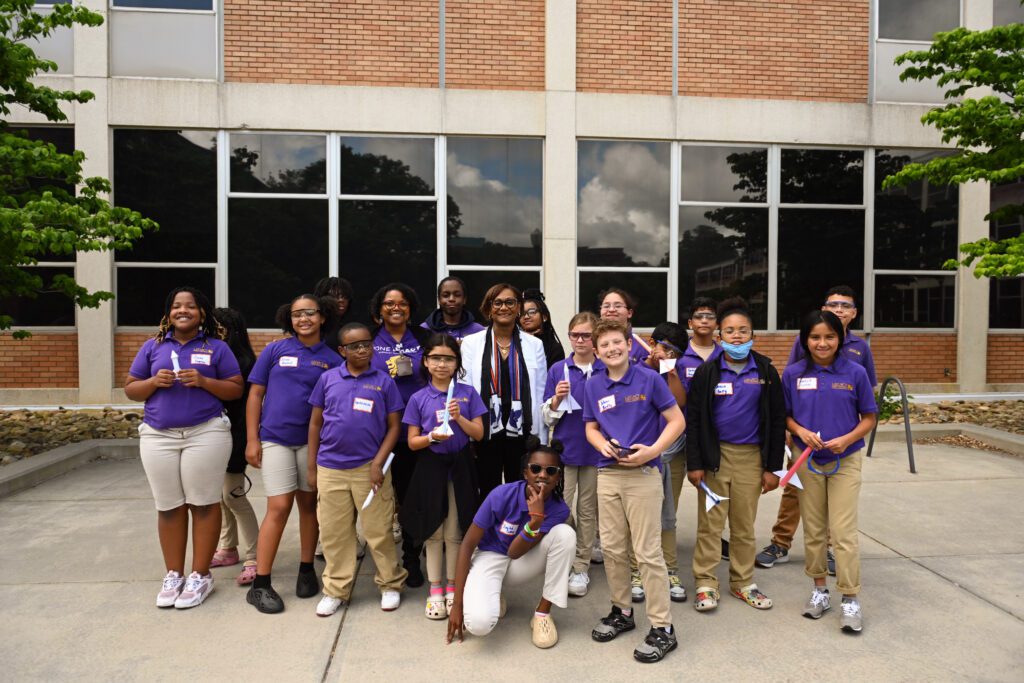 A group of elementary-age students all wearing purple Clemson shirts, some wearing lab glasses and masks, surrounds a Black woman wearing a white blazer and dark-rimmed glasses. They are standing outside and academic building.