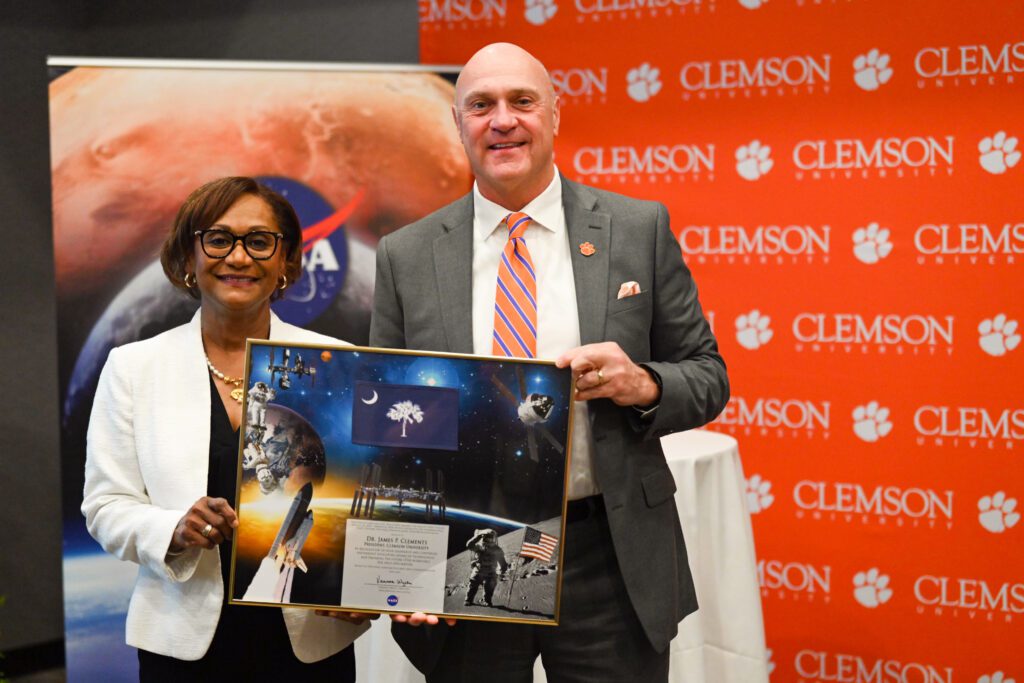 A black woman wearing a white blazer and glasses stands next to a white man wearing a grey suit and orange tie. They are holding a colorful document depicting outer space.
