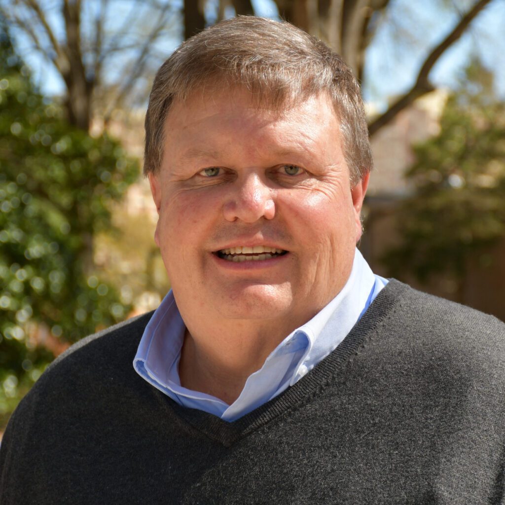 A smiling man wearing a gray sweater and blue Oxford shirt underneath