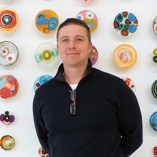 A smiling man wearing a blue sweater with folded glasses tucked into the zipper standing in front of a color array of plates on a wall