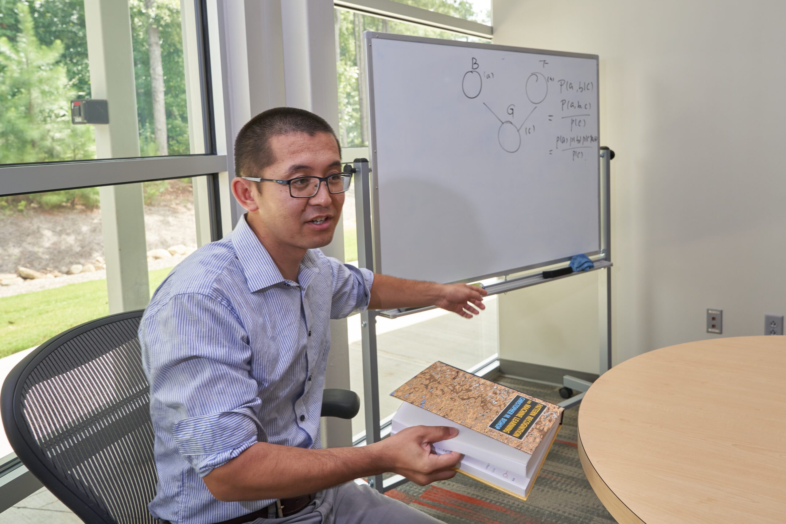 A man wearing glasses points at a whiteboard while holding a book