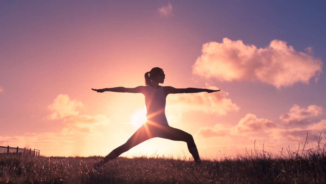 A woman does the "warrior" yoga pose in front of a sunset.